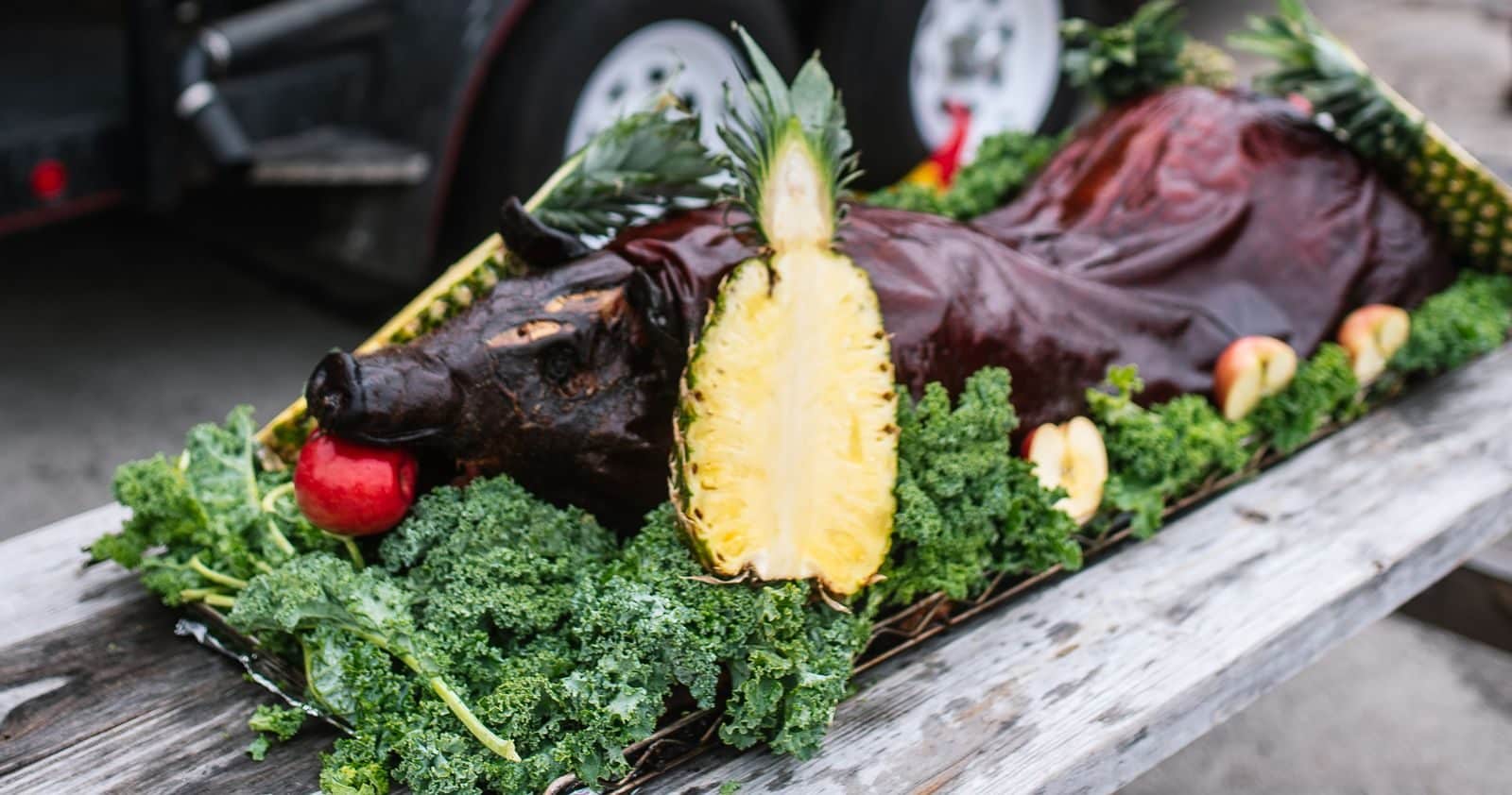 Whole Hog Roast 3 A whole roasted hog on kale and pineapple, set on a picnic table in front of the Salt + Smoke smoker trailer