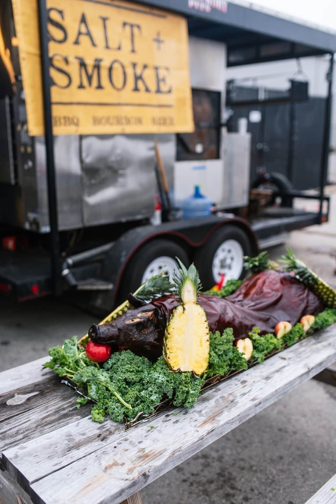 Whole Hog Roast 2 A whole roasted hog on kale and pineapple, set on a picnic table in front of the Salt + Smoke smoker trailer