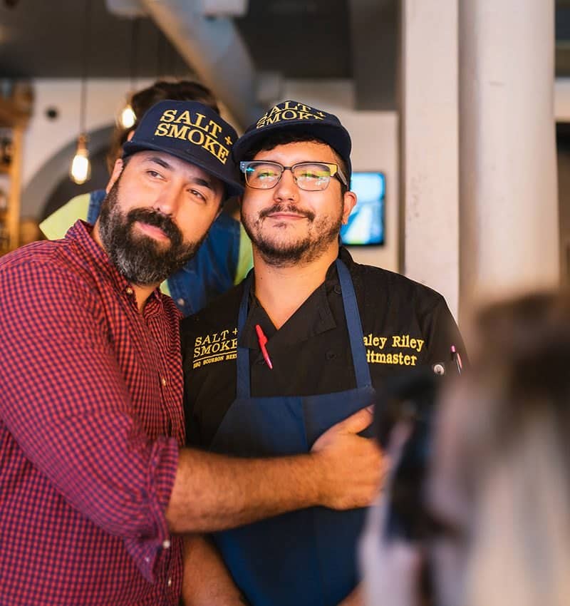 Owner Tom Schmidt with his arm around pitmaster Haley Riley, both in Salt + Smoke hats