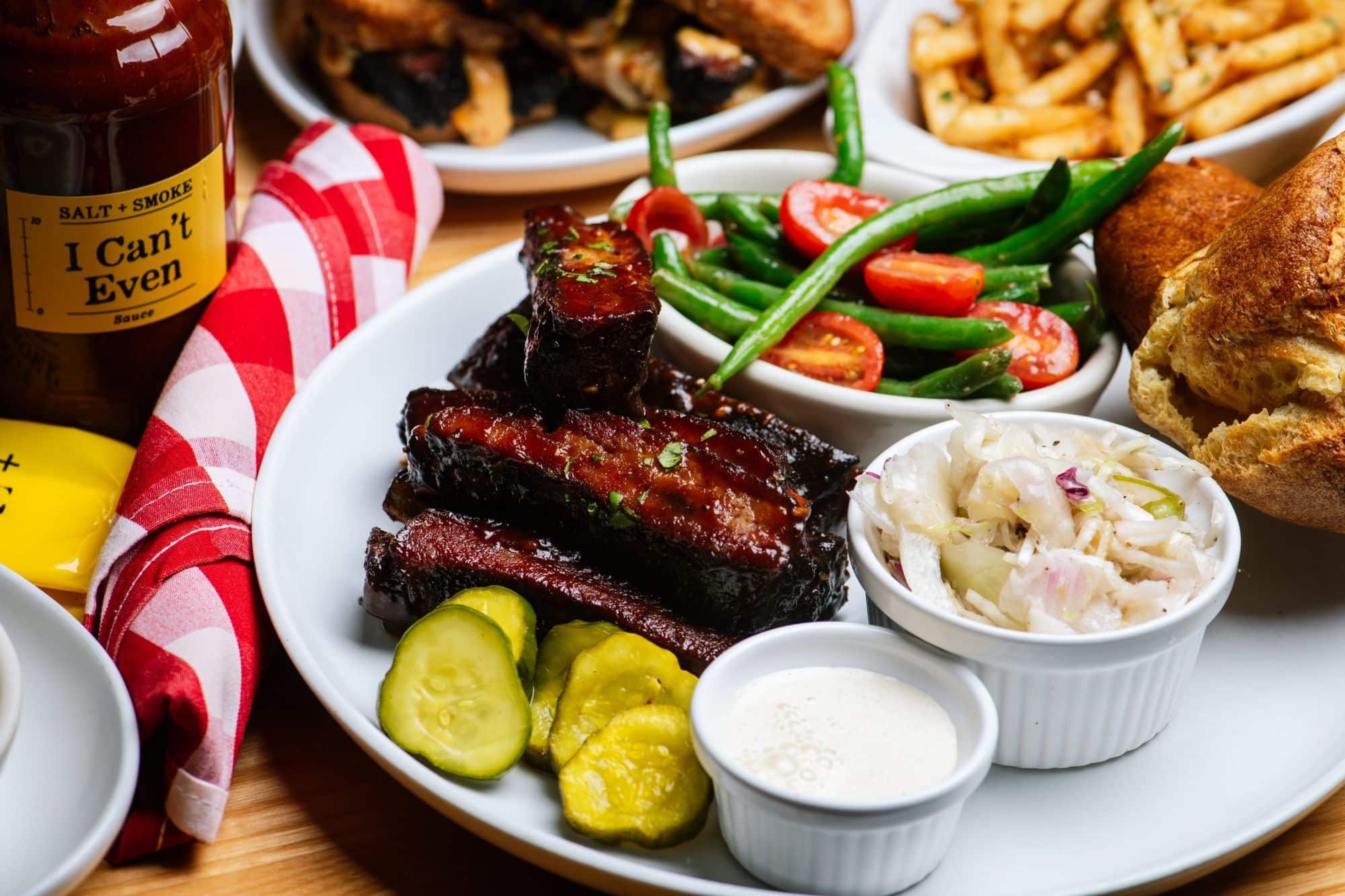 Home 1 Sticky trashed ribs with pickles, coleslaw, green bean salad and a popover, next to a bottle of I Can’t Even sauce